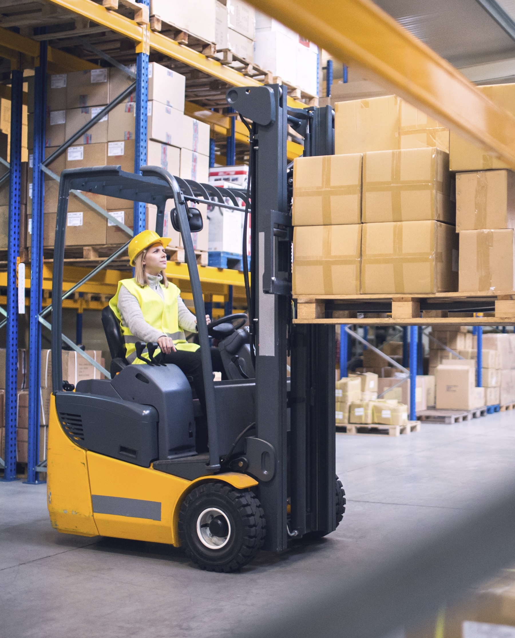 Besku-Logistics-logistiikan-tyontekijan-vuokraus-henkilostovuokraus-uusimaa-paakaupunkiseutu-Helsinki-Vantaa-Espoo-logistiikkavarasto-trukkikuski-tyovoimavuokraus Nuori nainen nostaa trukilla paketteja hyllyyn logistiikan varastohallissa.